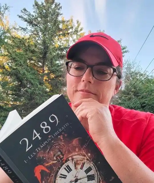 Une femme (l'auteure) en T-shirt rouge et casquette rouge lit "2489 - Les Temps Libératis" dans un décor de nature avec des arbres et une horloge sur la couverture.