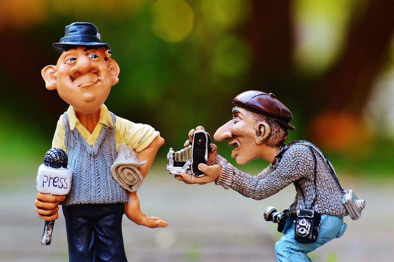 Figurines humoristiques représentant deux journalistes : l’un tient un micro avec l’étiquette "Press" et un journal sous le bras, tandis que l’autre prend une photo avec un vieil appareil photo, portant une caméra et un appareil photo autour du cou.